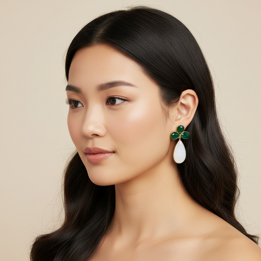 A close-up portrait of a woman with long dark hair wearing a distinctive drop earring. The earring features a four-petal flower design in polished green agate with a teardrop-shaped white chalcedony stone hanging below, set against a solid beige background.