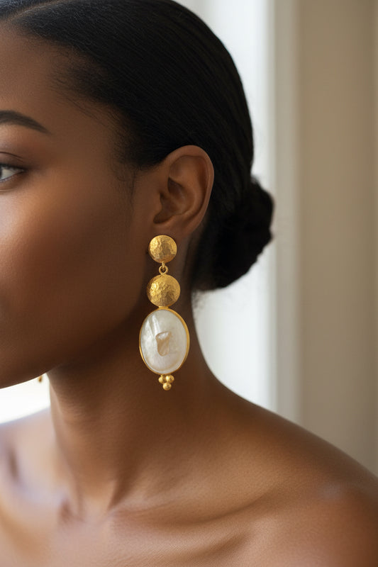 A pair of gold-toned drop earrings with textured round gold beads, oval white mother of pearl pendants, and small gold bead drops, displayed on a dark reddish-brown wooden surface.