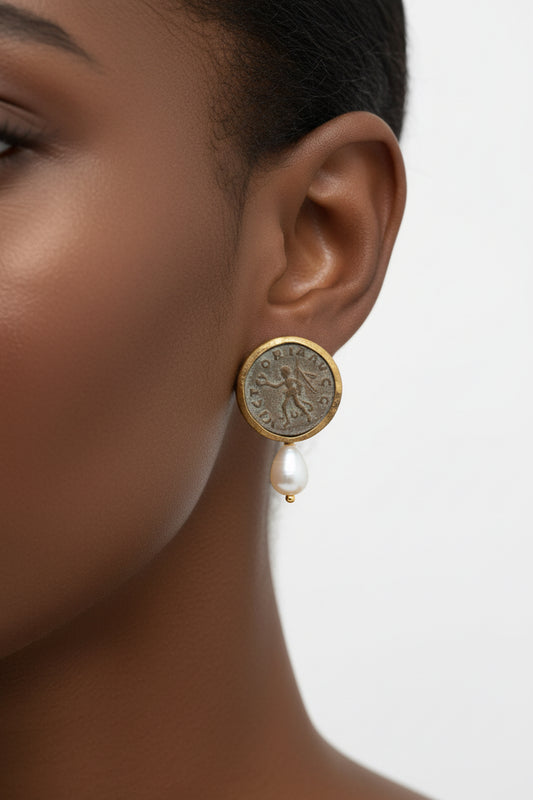 A small gold-toned earring featuring an antique-style coin face with a dangling pearl, resting on a dark reddish-brown wooden surface.