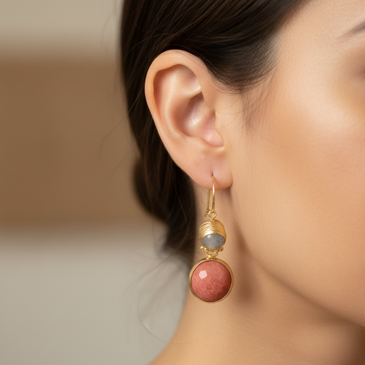A close-up of a model's ear wearing a gold drop earring with a round pink agate and a smaller round cat eye stone.