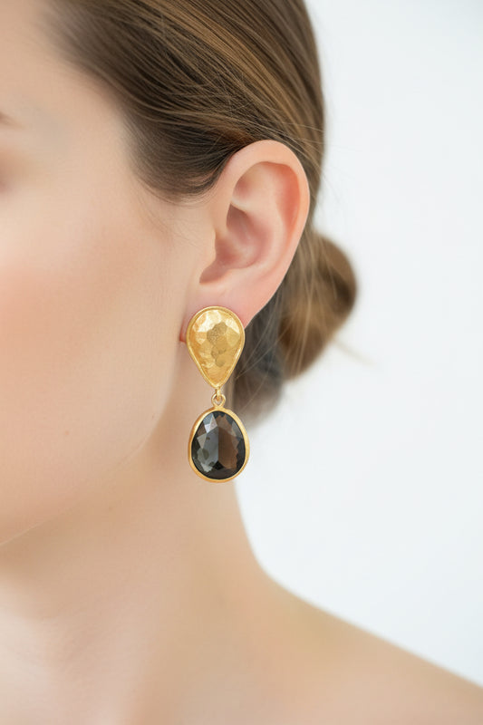 A pair of gold-toned clip-on earrings with teardrop-shaped gold tops and large oval dark gray labradorite faceted stones, resting on a dark red wooden surface.